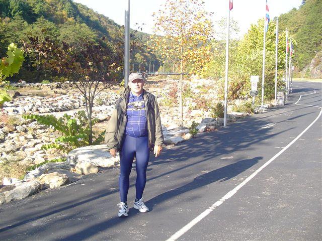 A person standing on a paved road surrounded by trees and rocks, with colorful autumn foliage. The individual is wearing a baseball cap, a jacket over a long-sleeved shirt, and form-fitting athletic pants. Flags are visible in the background, and the setting appears to be a natural landscape. Tanasi Trail System mountain bike trail.