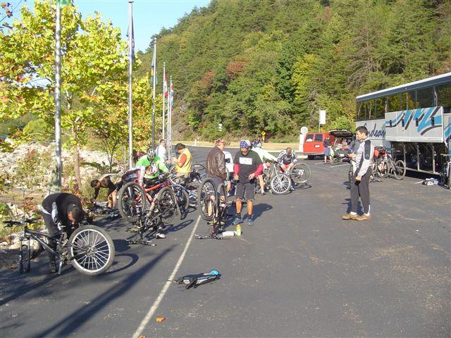 A group of cyclists working on their mountain bikes along a roadside, surrounded by trees and flags. Some individuals are repairing their bikes while others are observing. A bus is parked nearby. The scene captures an outdoor cycling event or gathering. Tanasi Trail System mountain bike trail.