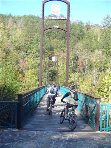 Two mountain bikers ride across a wooden bridge adorned with a sign that reads "Centennial Olympic 1996," surrounded by lush greenery and trees in various autumn colors. The scene captures a serene outdoor activity in a nature setting. Tanasi Trail System mountain bike trail.