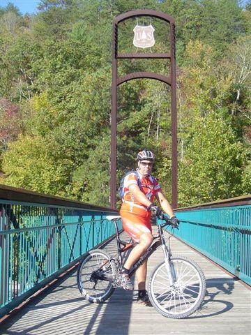 A cyclist in a colorful outfit stands on a bridge, posing with a mountain bike. The background features lush green trees and a sign marking the area. The bridge has a distinctive arch and a shield emblem at the top, suggesting it is a notable hiking or biking location. Tanasi Trail System mountain bike trail.