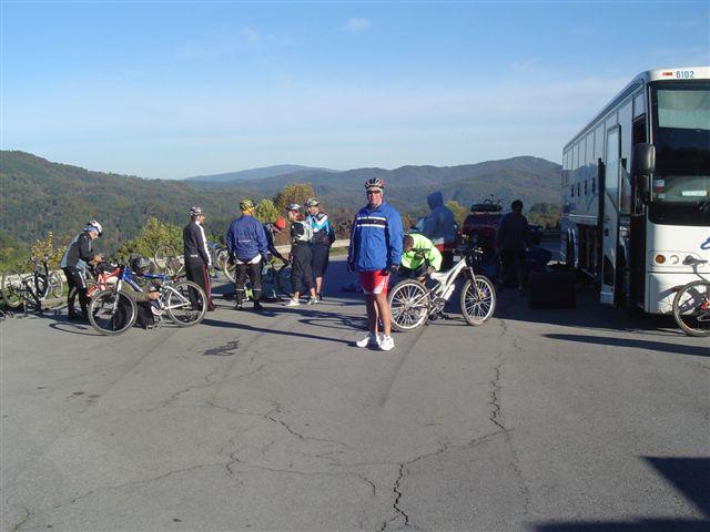 A group of cyclists preparing for a ride, with some gathered around bicycles and others near a bus. The setting features a scenic view of mountains in the background under a clear blue sky. The cyclists are dressed in athletic wear, and a few are adjusting their gear. Tanasi Trail System mountain bike trail.