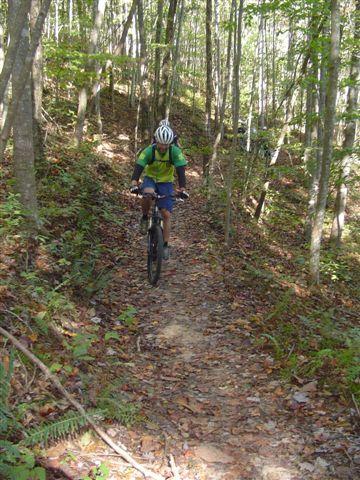 A person wearing a helmet and cycling gear rides a mountain bike along a narrow dirt trail in a forested area, surrounded by trees and foliage. Tanasi Trail System mountain bike trail.