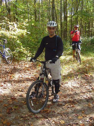 A male mountain biker in a helmet and protective gear stands beside his bicycle on a forest trail surrounded by trees and autumn foliage. Another cyclist is visible in the background, adjusting their camera. Tanasi Trail System mountain bike trail.