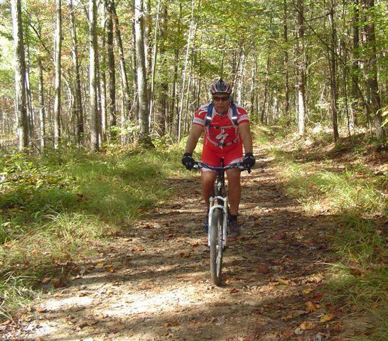 A man riding a mountain bike on a forest trail surrounded by trees, wearing a red cycling jersey and helmet, with sunlight filtering through the leaves. Tanasi Trail System mountain bike trail.
