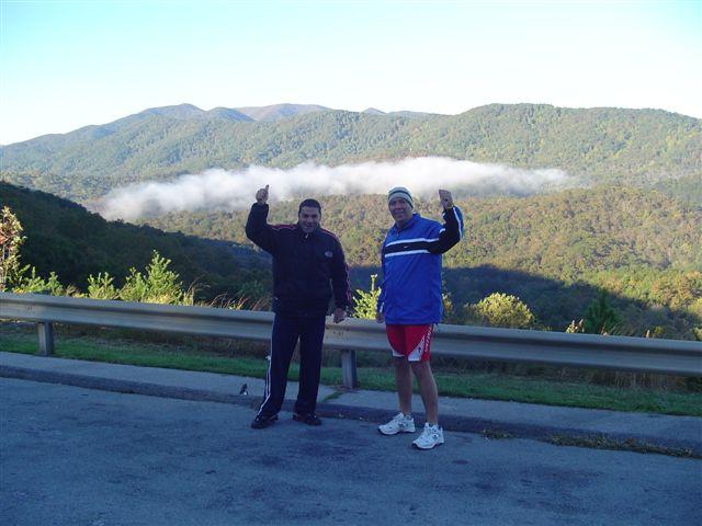 Two men standing on a roadside with their arms raised, smiling at the camera. They are in front of a picturesque mountainous landscape covered in greenery and morning mist. The sun is shining, and the sky is clear blue, indicating a beautiful day. Tanasi Trail System mountain bike trail.