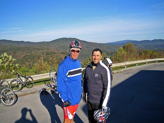 Two male cyclists posing for a photo with a scenic mountain background. They are wearing cycling gear and helmets, with bicycles parked nearby. The landscape features rolling hills and trees, showcasing a clear blue sky and autumn foliage. Tanasi Trail System mountain bike trail.