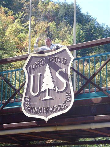 A person stands atop a bridge, holding up a large sign for the U.S. Forest Service, which features a tree emblem and the text "DEPARTMENT OF AGRICULTURE." The background shows trees with autumn foliage. Tanasi Trail System mountain bike trail.