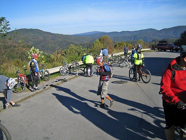 A group of cyclists gathered on a road surrounded by mountains, with several bicycles lying on the ground. Some cyclists are working on their bikes, while others are standing or walking around. The background features a picturesque view of rolling hills and clear skies. Tanasi Trail System mountain bike trail.