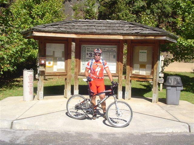 A cyclist in red and white biking gear stands next to a mountain bike at a trailhead information kiosk surrounded by greenery. The kiosk features various informational postings and a trash can nearby. Tanasi Trail System mountain bike trail.