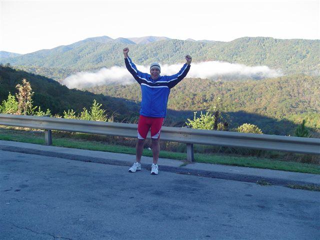 A person stands outdoors with arms raised in celebration, wearing a blue and white jacket, red shorts, and sneakers. In the background, there are rolling mountains with a layer of fog hovering between them and clear blue skies above. The scene conveys a sense of achievement and joy in a natural setting. Tanasi Trail System mountain bike trail.