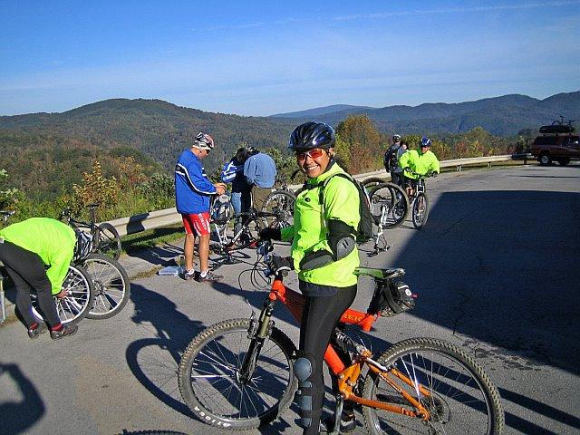 A group of cyclists is gathered on a scenic mountain overlook, preparing their bikes and taking a break. One woman in the foreground is smiling and wearing a bright green jacket and helmet while standing next to her orange mountain bike. The others are engaged in adjusting their bikes, with rolling hills and blue skies in the background. Tanasi Trail System mountain bike trail.