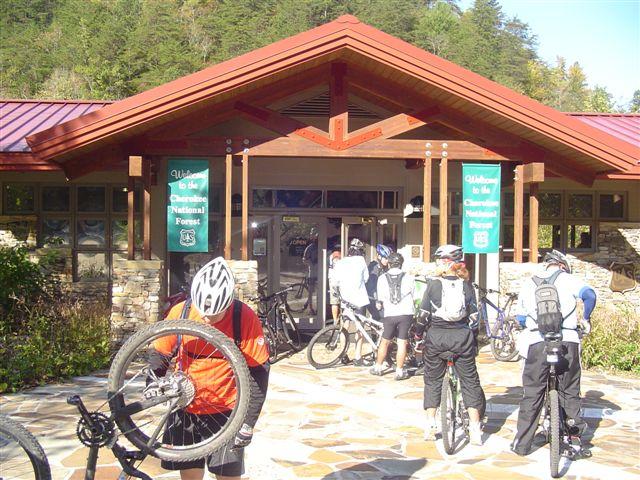 A group of mountain bikers gather outside a visitor center at the Cheoah National Forest, featuring a wooden structure with a red roof. The entrance has welcoming signs, and bikers are seen preparing their bikes on a stone pathway surrounded by trees. Tanasi Trail System mountain bike trail.