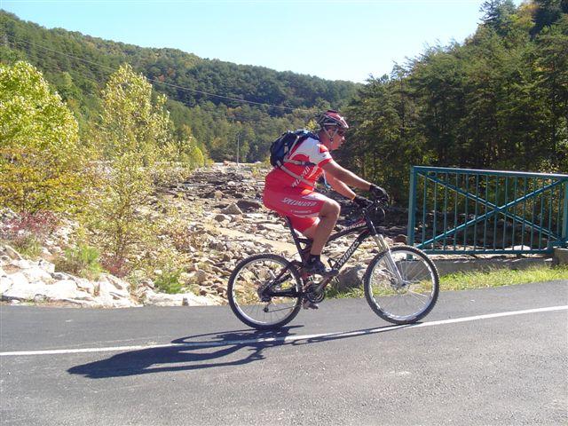 A cyclist in a red and white jersey rides a mountain bike along a scenic road surrounded by trees and hills. In the background, there is a rocky area and a green gate. The sun is shining, indicating a clear day. Tanasi Trail System mountain bike trail.