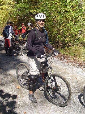 A man in cycling gear smiles while standing next to a mountain bike on a gravel trail. He wears a helmet, knee pads, and a backpack, surrounded by other cyclists in the background. The setting features lush green trees, indicating a forested area suitable for biking. Tanasi Trail System mountain bike trail.