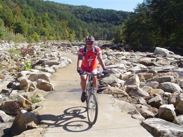 A mountain biker riding on a rocky trail surrounded by trees and hills. The cyclist, wearing a red jersey and a helmet, is navigating a path paved with stones and bordered by large boulders. Bright sunlight illuminates the scene, highlighting the vibrant colors of the autumn foliage in the background. Tanasi Trail System mountain bike trail.