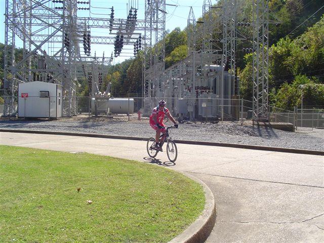 A cyclist in red gear rides along a paved path near a power station, surrounded by electrical equipment and greenery in the background. Tanasi Trail System mountain bike trail.