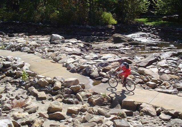 A cyclist riding on a rocky path beside a stream, surrounded by large boulders and green trees. The scene is illuminated by sunlight, highlighting the natural landscape. Tanasi Trail System mountain bike trail.