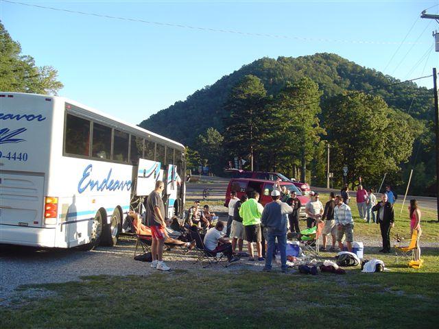 A group of people gathered around a bus labeled "Endeavor" in a scenic outdoor setting, with green mountains in the background. Some individuals are sitting on folding chairs, while others are standing and engaged in conversation. The scene conveys a sense of camaraderie and leisure among travelers. Tanasi Trail System mountain bike trail.