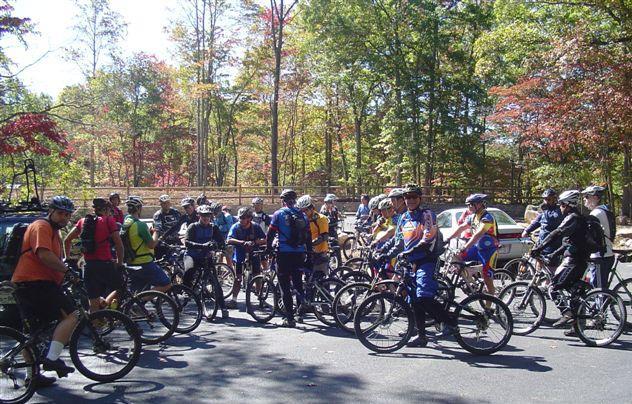 A group of cyclists gathered on mountain bikes in a wooded area, surrounded by autumn foliage. The scene depicts a diverse group of individuals wearing cycling gear, with some standing beside their bikes and others seated, ready for a ride. The background features trees displaying fall colors, and a vehicle can be seen parked nearby. Chilhowee trail system mountain bike trail.