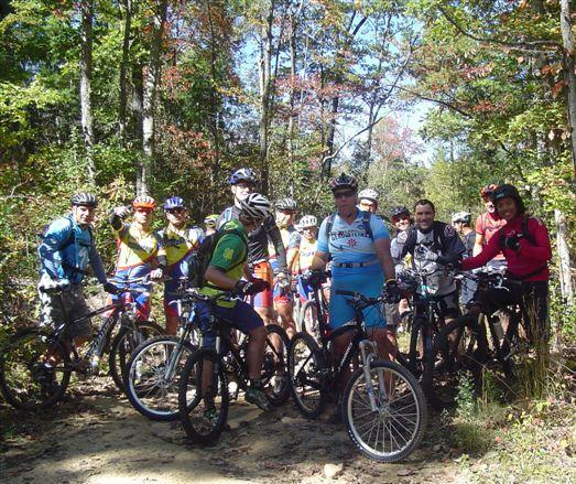 A group of mountain bikers, dressed in colorful cycling gear and helmets, pose together on a dirt path surrounded by trees in a forested area. They appear to be in a cheerful mood and are gathered near their bikes, ready for a ride or a break. The photo captures a bright, sunny day with clear blue skies and hints of autumn foliage in the background. Chilhowee trail system mountain bike trail.