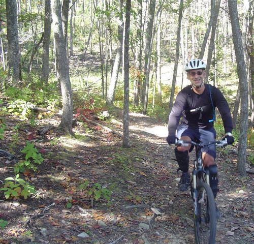 A mountain biker riding along a forest trail during daylight, surrounded by trees and colorful autumn leaves. The cyclist is wearing a helmet and a black long-sleeve shirt, smiling as he navigates the terrain. Chilhowee trail system mountain bike trail.