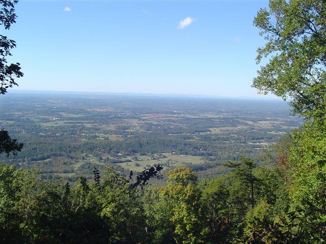 A scenic view of a lush green valley surrounded by distant mountains under a clear blue sky, with a few scattered clouds. The foreground features trees and foliage, while the expansive landscape below includes fields and small patches of farmland. Chilhowee trail system mountain bike trail.