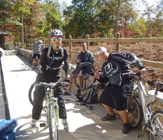 Three mountain bikers are gathered at a trailhead on a sunny day. One rider, wearing a helmet and sunglasses, stands next to a green mountain bike, smiling at the camera. Another rider is seated nearby, working on a bike tire, while a third rider leans in, giving a peace sign. The background features colorful autumn foliage and a wooden fence. Chilhowee trail system mountain bike trail.