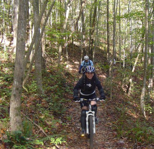 Two mountain bikers navigating a dirt trail through a lush forest, with trees on either side and sunlight filtering through the leaves. One rider is in the foreground wearing a dark jacket and helmet, while the other is visible in the background. Chilhowee trail system mountain bike trail.