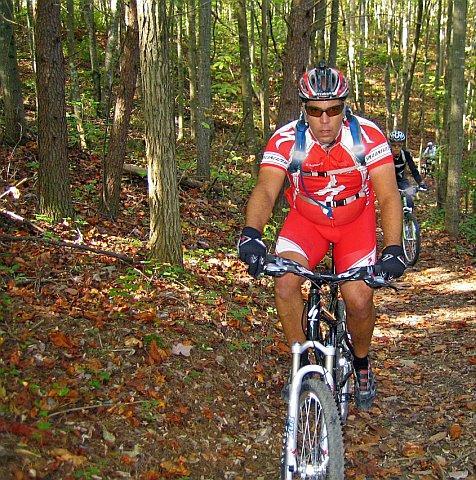 A male mountain biker wearing a red and white cycling outfit rides along a forest trail surrounded by trees and autumn foliage. His focused expression and athletic posture suggest a sense of determination as he navigates the uneven terrain. In the background, another cyclist is visible, also biking on the trail. Chilhowee trail system mountain bike trail.