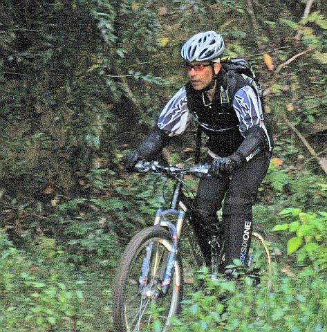 A cyclist wearing a helmet and protective gear rides a mountain bike along a wooded trail, surrounded by greenery. Chilhowee trail system mountain bike trail.