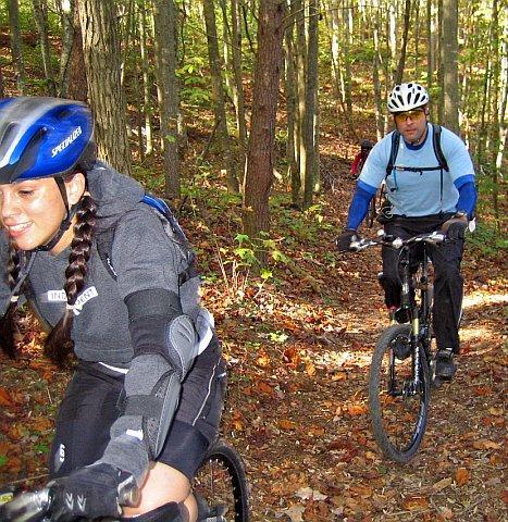 A girl and a man biking along a narrow dirt trail through a forest. The girl is wearing a blue helmet and protective gear, smiling as she rides ahead. The man is trailing behind, wearing a helmet and light blue cycling attire, focused on the path. The surroundings are filled with trees and fallen leaves, showcasing an autumn setting. Chilhowee trail system mountain bike trail.