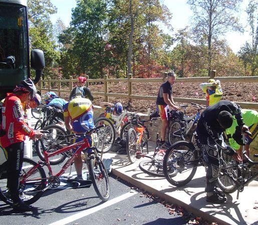 Groups of cyclists in colorful jerseys are gathered around their bicycles, working on repairs or adjustments. The scene takes place outdoors in a wooded area with trees displaying autumn foliage. A bus is parked nearby, and the cyclists are focused on their tasks, indicating a break or preparation during a biking event. Chilhowee trail system mountain bike trail.