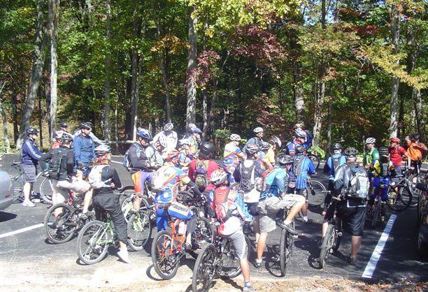 A large group of mountain bikers gathered in a wooded area, preparing for a ride. The participants, wearing helmets and cycling gear, are standing next to their bikes, with trees displaying autumn foliage in the background. Chilhowee trail system mountain bike trail.