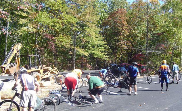 A group of people repairing bicycles on a paved road surrounded by trees with autumn foliage. Some individuals are bent over their bikes, while others are standing; a construction vehicle is visible in the background. Chilhowee trail system mountain bike trail.