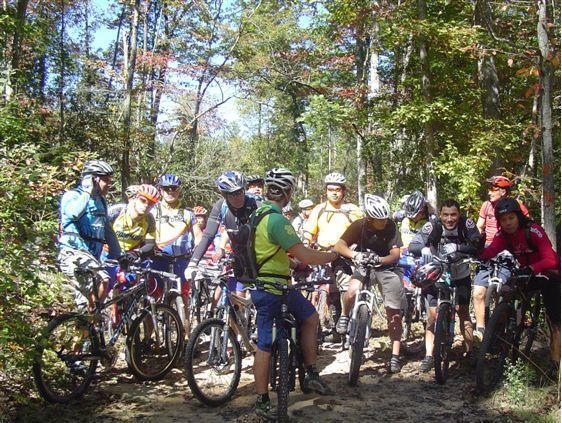 Group of mountain bikers gathered on a dirt trail in a wooded area, wearing helmets and cycling gear. The scene captures a mix of cyclists stopped during a ride, surrounded by trees with signs of autumn foliage. Chilhowee trail system mountain bike trail.