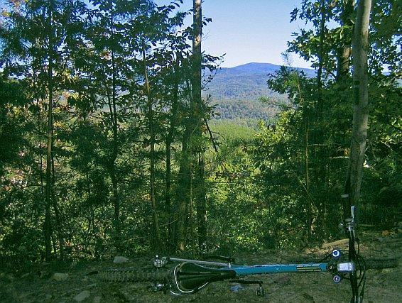 A scenic view of a mountainous landscape framed by trees, with a blue mountain bike laying on the ground in the foreground. The sky is clear, and the greenery of the forest contrasts with the distant mountains. Chilhowee trail system mountain bike trail.