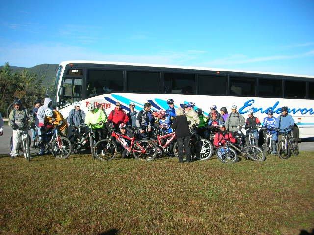 A group of cyclists, wearing colorful cycling gear and helmets, gathers in front of a large bus parked on grass. The bus is labeled "Endeavors," and the background features a clear blue sky and distant mountains. The cyclists are posed with their bicycles, showing a mix of excitement and camaraderie. Tanasi Trail System mountain bike trail.