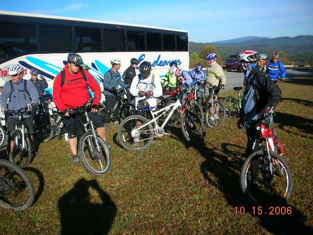 A group of mountain bikers in helmets gathered around their bikes in front of a bus on a grassy area, with mountains visible in the background under a clear blue sky. Tanasi Trail System mountain bike trail.