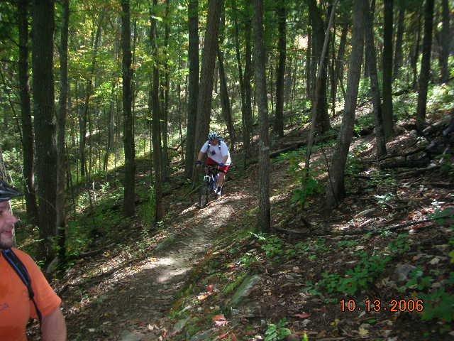 A mountain biker riding along a narrow trail in a lush green forest, with trees surrounding the path. In the foreground, another person wearing an orange shirt is observing. The scene captures a sunny day in the woods, highlighting the beauty of nature. Tanasi Trail System mountain bike trail.