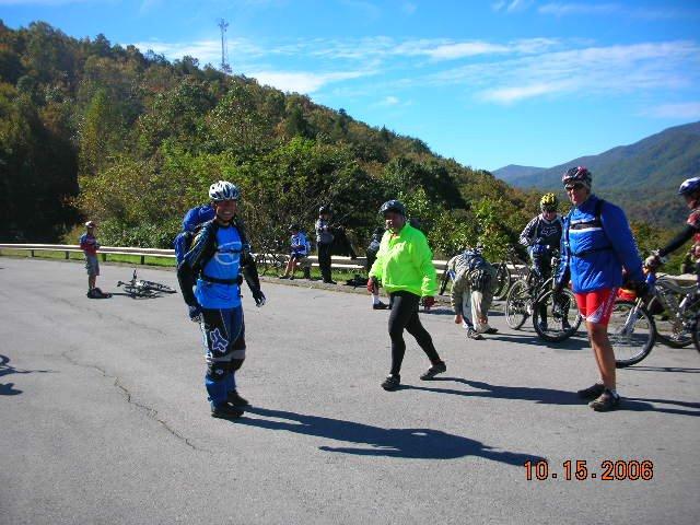 A group of cyclists wearing bright apparel stand on a paved area surrounded by greenery and mountains. Some riders are interacting and smiling, while others are preparing their bikes. The scene is set on a sunny day with clear blue skies, showcasing an outdoor cycling event. Tanasi Trail System mountain bike trail.