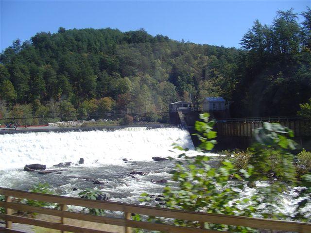 A scenic view of a waterfall cascading over a dam, surrounded by greenery and trees. The water flows vigorously over the edge, with rocky formations visible in the foreground. In the background, a small structure is nestled among the trees, enhancing the natural landscape. Blue skies provide a bright backdrop to the scene. Tanasi Trail System mountain bike trail.