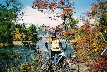 A person in cycling gear stands next to a mountain bike, surrounded by autumn foliage and trees. In the background, a river flows under a partly cloudy blue sky. Tsali Left Loop mountain bike trail.