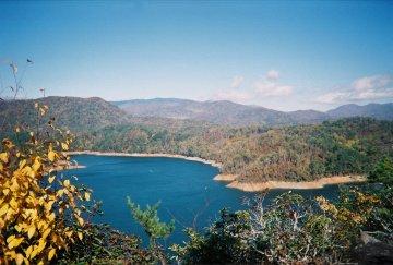 A scenic view of a tranquil lake surrounded by lush, rolling hills and mountains. The water reflects the clear sky, and the shoreline is framed by trees exhibiting vibrant autumn foliage. The landscape conveys a sense of peace and natural beauty. Tsali Left Loop mountain bike trail.