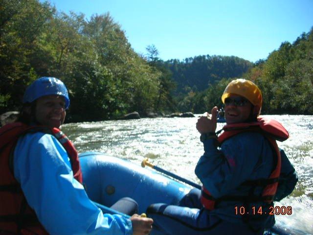 Two people are sitting in a blue inflatable raft on a river surrounded by green trees and hills. Both are wearing helmets and life jackets. One person is giving a thumbs up, smiling, while the other looks towards the camera. The scene is bright, suggesting a pleasant day of rafting. Tanasi Trail System mountain bike trail.