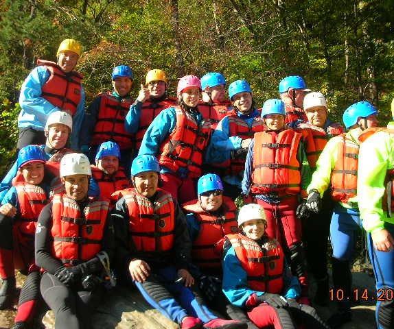 A group of people in colorful rafting gear, including bright orange life vests and helmets, posing together outdoors. They are gathered on a rocky area surrounded by trees, smiling and looking excited, indicating a fun outdoor adventure. The scene captures a sense of camaraderie and adventure. Tanasi Trail System mountain bike trail.