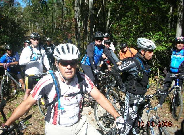 A group of mountain bikers wearing helmets and cycling gear, gathered on a wooded trail. The scene captures several individuals on their bikes, with a focus on one rider in the foreground. The image conveys a sense of camaraderie among the cyclists, enjoying a day outdoors in nature. Tanasi Trail System mountain bike trail.