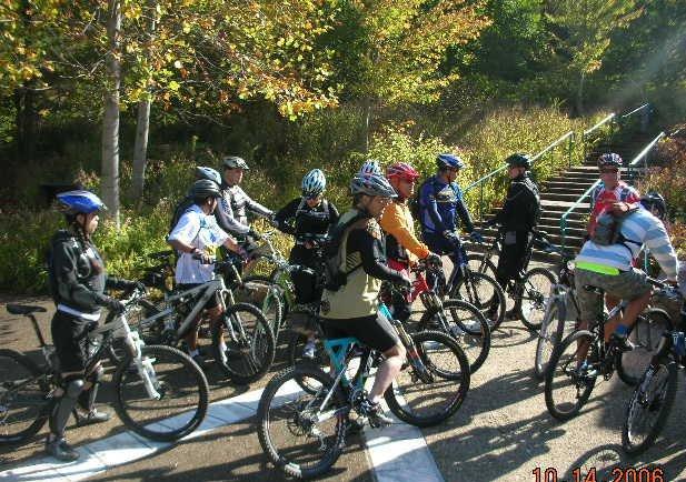 A group of mountain bikers gathered on a sunny day, wearing cycling helmets and gear, with various types of bicycles. They are positioned on a path with greenery and trees in the background, preparing for a ride. The atmosphere is lively and engaged, suggesting camaraderie and adventure among the cyclists. Tanasi Trail System mountain bike trail.