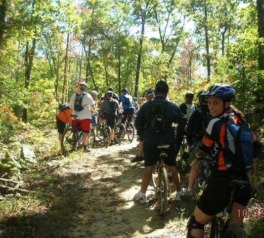 A group of people biking on a trail surrounded by trees in a wooded area. They are wearing helmets and cycling gear, with some participants standing beside their bikes while others are seated. The scene captures a sunny day with vibrant autumn foliage. Tanasi Trail System mountain bike trail.