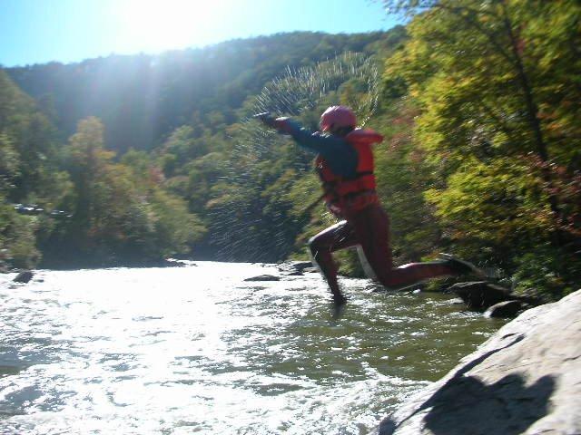 A person wearing a red life jacket and helmet leaps into a river, splashing water around them. The scene is set in a scenic location with lush green hills in the background, illuminated by bright sunlight. Tanasi Trail System mountain bike trail.