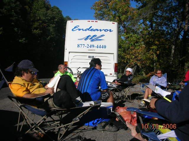 A group of people sitting in folding chairs in a parking area, with a large white bus labeled "Endeavor" in the background. The scene is set outdoors with trees showing autumn foliage. Some individuals are engaged in conversation, while others are reading or writing. It appears to be a casual gathering or break during an outdoor activity. Tanasi Trail System mountain bike trail.